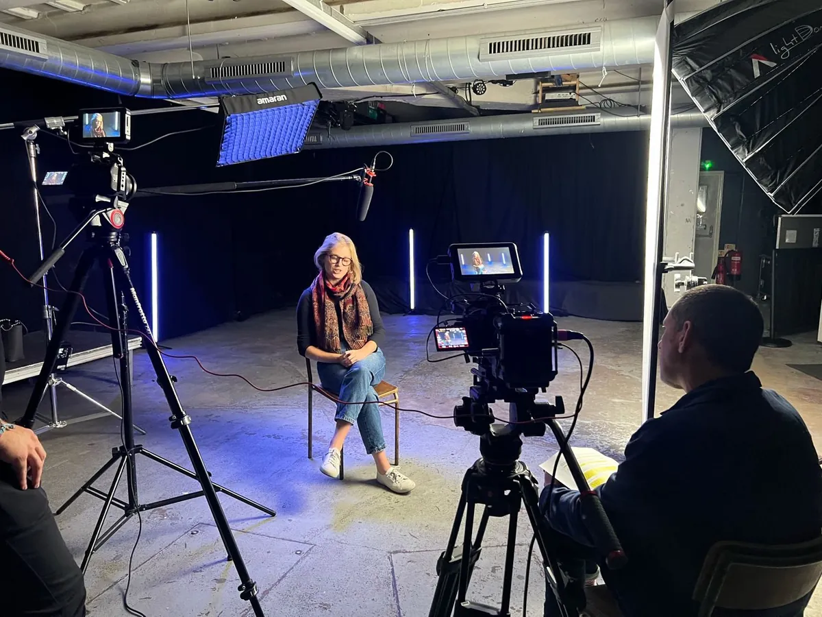 A contributor mid-interview in a dark studio, framed by two cameras, a boom microphone and cool blue tube lighting